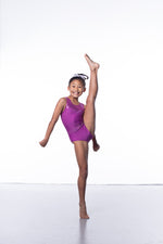 Photo of a 10 year old girl standing infront of a white background. Wearing a short sleeve plum coloured gymnastics leotard. The top of the leotard is a shiny plum fabric, and the lower is a matte plum fabric. The matte fabric has a heavy amount of crystal detailing, to make it very sparkly. 