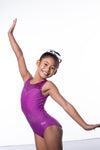 Photo of a 10 year old girl standing infront of a white background. Wearing a short sleeve plum coloured gymnastics leotard. The top of the leotard is a shiny plum fabric, and the lower is a matte plum fabric. The matte fabric has a heavy amount of crystal detailing, to make it very sparkly. 