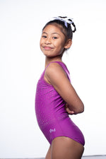 Photo of a 10 year old girl standing infront of a white background. Wearing a short sleeve plum coloured gymnastics leotard. The top of the leotard is a shiny plum fabric, and the lower is a matte plum fabric. The matte fabric has a heavy amount of crystal detailing, to make it very sparkly. 