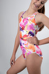 Girl standing infront of a grey background, wearing a bright floral gymnastics leotard that is a mix of purple, orange, white & pink in colour. The back of the leotard has a triangle cut out, with straps that pull through the cut out, creating a stuning back feature. 