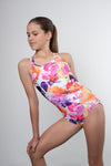 Girl standing infront of a grey background, wearing a bright floral gymnastics leotard that is a mix of purple, orange, white & pink in colour. The back of the leotard has a triangle cut out, with straps that pull through the cut out, creating a stuning back feature. 