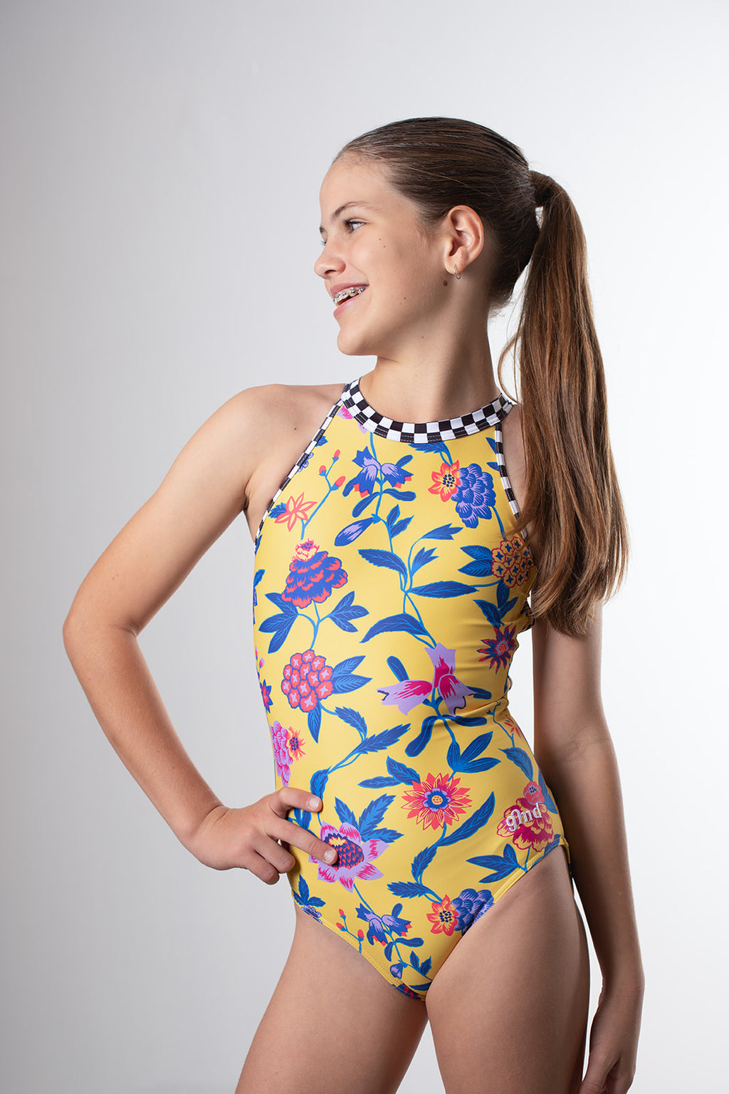 Girl standing infront of a pink backdrop, wearing a yellow floral gymnastics leotard. The back of the leotard features black & white checkered criss cross strap details for contrast.