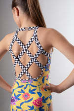 Girl standing infront of a pink backdrop, wearing a yellow floral gymnastics leotard. The back of the leotard features black & white checkered criss cross strap details for contrast.
