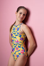 Girl standing infront of a pink backdrop, wearing a yellow floral gymnastics leotard. The back of the leotard features black & white checkered criss cross strap details for contrast.