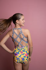 Girl standing infront of a pink backdrop, wearing a yellow floral gymnastics leotard. The back of the leotard features black & white checkered criss cross strap details for contrast.