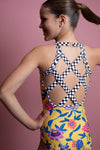 Girl standing infront of a pink backdrop, wearing a yellow floral gymnastics leotard. The back of the leotard features black & white checkered criss cross strap details for contrast.