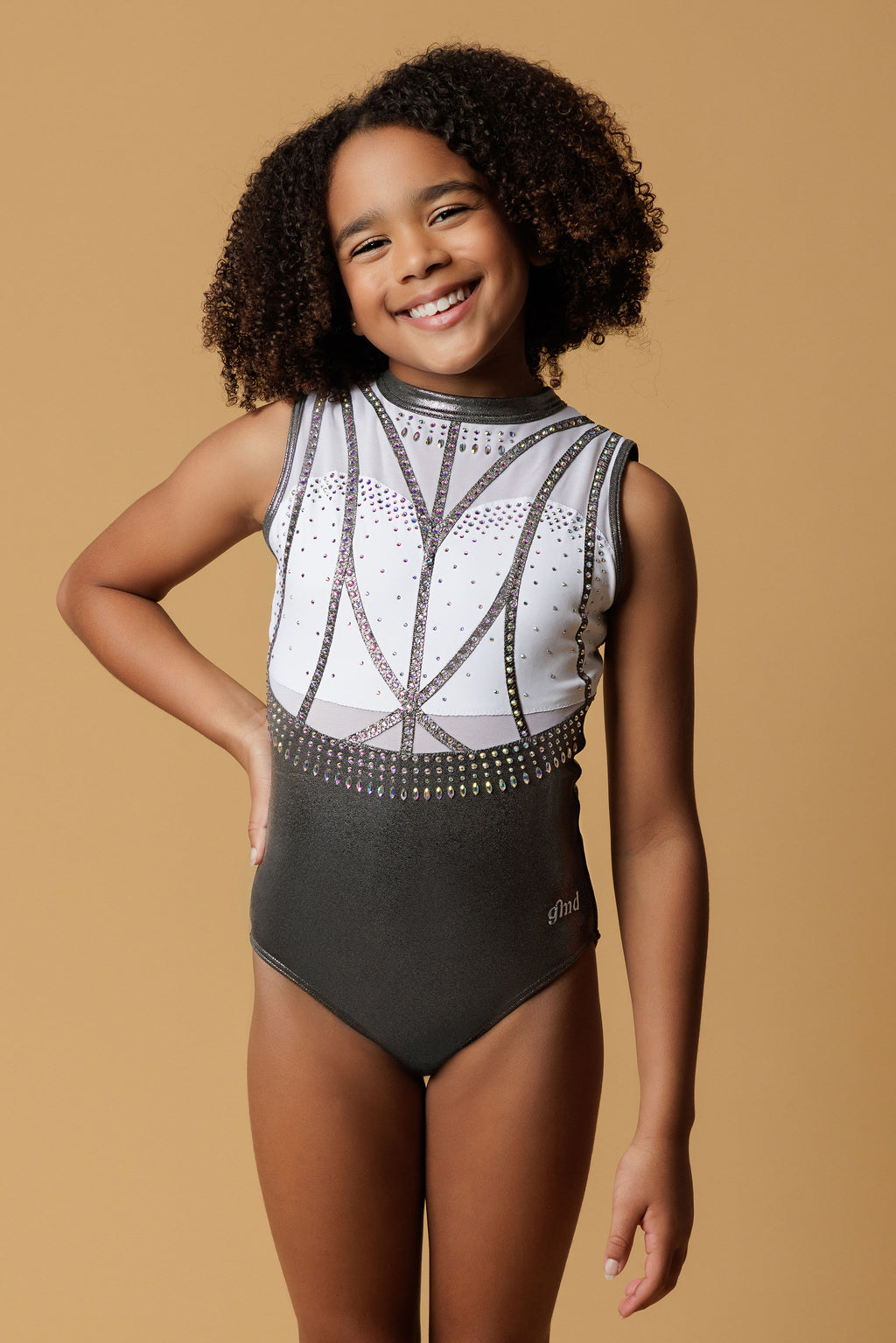 Girl staning infront of a beige background, wearing a white & gunmetal grey gymnastics leotard. The top half of the letoard is white and is not see through. It is encrusted with crystals and features gunmetal grey shiny applique detailing. The lower half is shiny gunmetal mystique fabric. Fully lined.