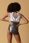 Girl staning infront of a beige background, wearing a white & gunmetal grey gymnastics leotard. The top half of the letoard is white and is not see through. It is encrusted with crystals and features gunmetal grey shiny applique detailing. The lower half is shiny gunmetal mystique fabric. Fully lined.