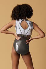 Girl staning infront of a beige background, wearing a white & gunmetal grey gymnastics leotard. The top half of the letoard is white and is not see through. It is encrusted with crystals and features gunmetal grey shiny applique detailing. The lower half is shiny gunmetal mystique fabric. Fully lined.