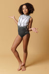 Girl staning infront of a beige background, wearing a white & gunmetal grey gymnastics leotard. The top half of the letoard is white and is not see through. It is encrusted with crystals and features gunmetal grey shiny applique detailing. The lower half is shiny gunmetal mystique fabric. Fully lined.