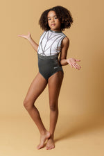 Girl staning infront of a beige background, wearing a white & gunmetal grey gymnastics leotard. The top half of the letoard is white and is not see through. It is encrusted with crystals and features gunmetal grey shiny applique detailing. The lower half is shiny gunmetal mystique fabric. Fully lined.