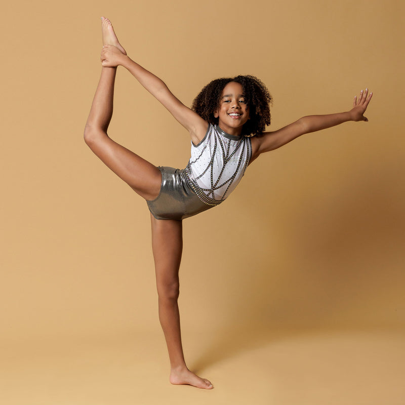 Girl staning infront of a beige background, wearing a white & gunmetal grey gymnastics leotard. The top half of the letoard is white and is not see through. It is encrusted with crystals and features gunmetal grey shiny applique detailing. The lower half is shiny gunmetal mystique fabric. Fully lined.