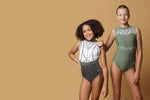 Girl staning infront of a beige background, wearing a white & gunmetal grey gymnastics leotard. The top half of the letoard is white and is not see through. It is encrusted with crystals and features gunmetal grey shiny applique detailing. The lower half is shiny gunmetal mystique fabric. Fully lined.