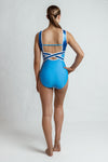 Photo of a girl standing infront of a white backdrop wearing a blue short sleeve gymnastics leotard. There are two tones of blue, one lighter and one darker shiny colour. There is white appliques and shiny diamantee detailing. The back has a cross over strap detailing.